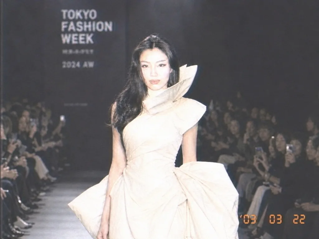 Keep this runway photo exactly as it is — the same woman, gown, pose, runway and audience — and only change its look to a still from a retro 2003 consumer camcorder: soft focus, washed-out colours, blown-out highlights and an orange date stamp in the corner.